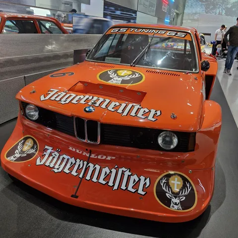 Front view of a bright orange 1979 BMW 320 Turbo Group 5 (E21) race car on display at the BMW Museum in Munich, featuring an ultra-wide body kit, large Jägermeister logos on the hood and massive front air dam, and a "GS TUNING GS" windshield banner.