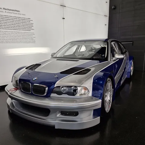 Three-quarters front view of a silver and blue BMW M3 GTR (E46) race car on display at the BMW Museum in Munich, featuring an aggressive front bumper, wide-body box fenders, hood vents, and a large rear race wing.