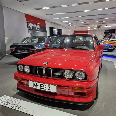 Front view of a bright red BMW M3 Sport Evolution (E30) on display at the BMW Museum in Munich, featuring quad round headlights, an M3 grille badge, iconic wide box fenders, and an adjustable front splitter with German license plate "M-ES 3".