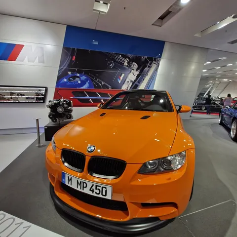 Front view of a bright Fire Orange BMW M3 GTS (E92) coupe on display at the BMW Museum in Munich, featuring an aggressive front bumper, dark kidney grilles, a prominent black front splitter, and license plate "M MP 450".
