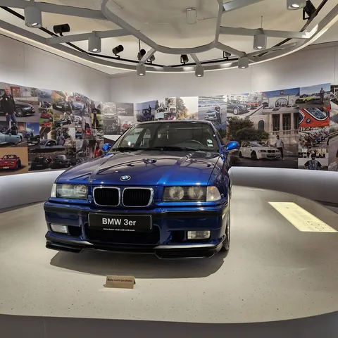 Front view of a dark metallic blue BMW 3 Series Coupe (E36) displayed in a circular hall at the BMW Museum in Munich, featuring an M-Technic front bumper with black splitters, clear glass dual headlights, and a wall of enthusiast photo collages in the background.